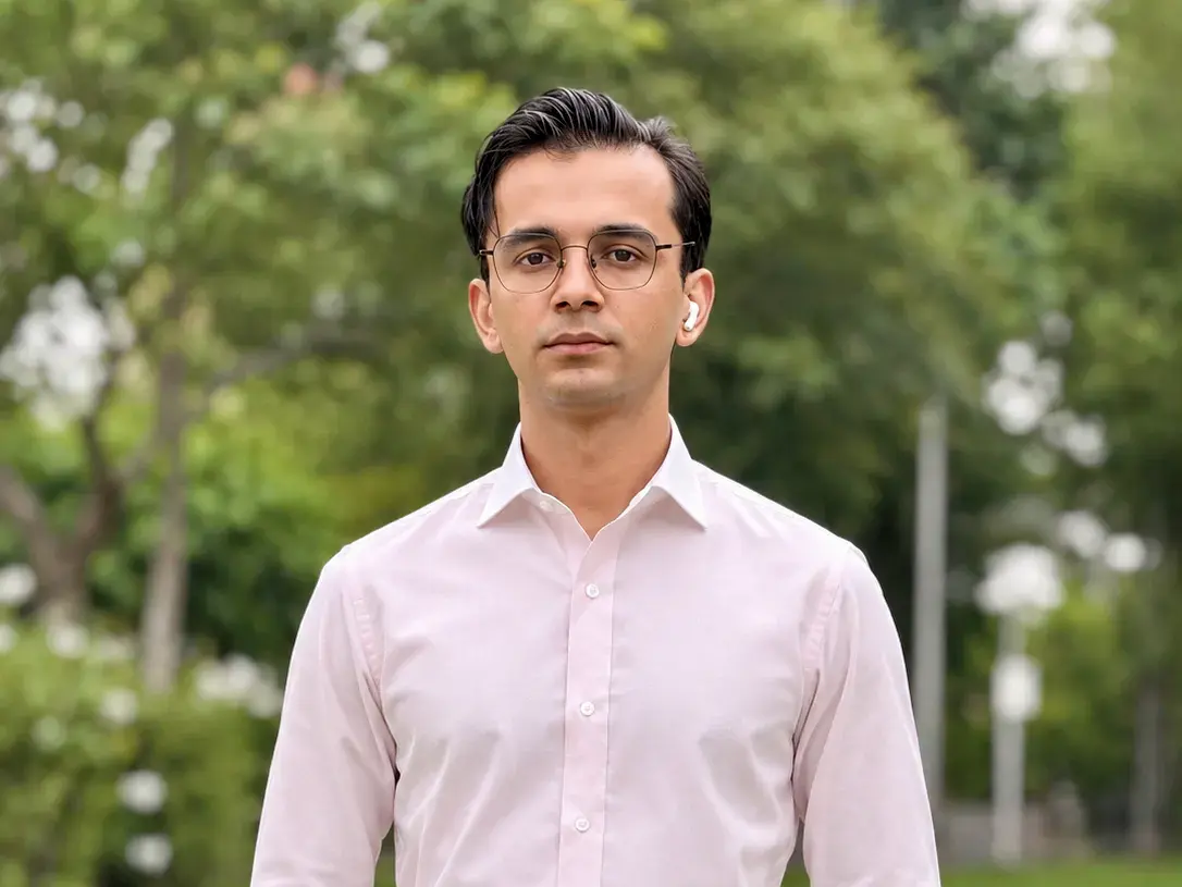 Man in a tailored light pink dress shirt and high-waisted grey trousers standing by a pond