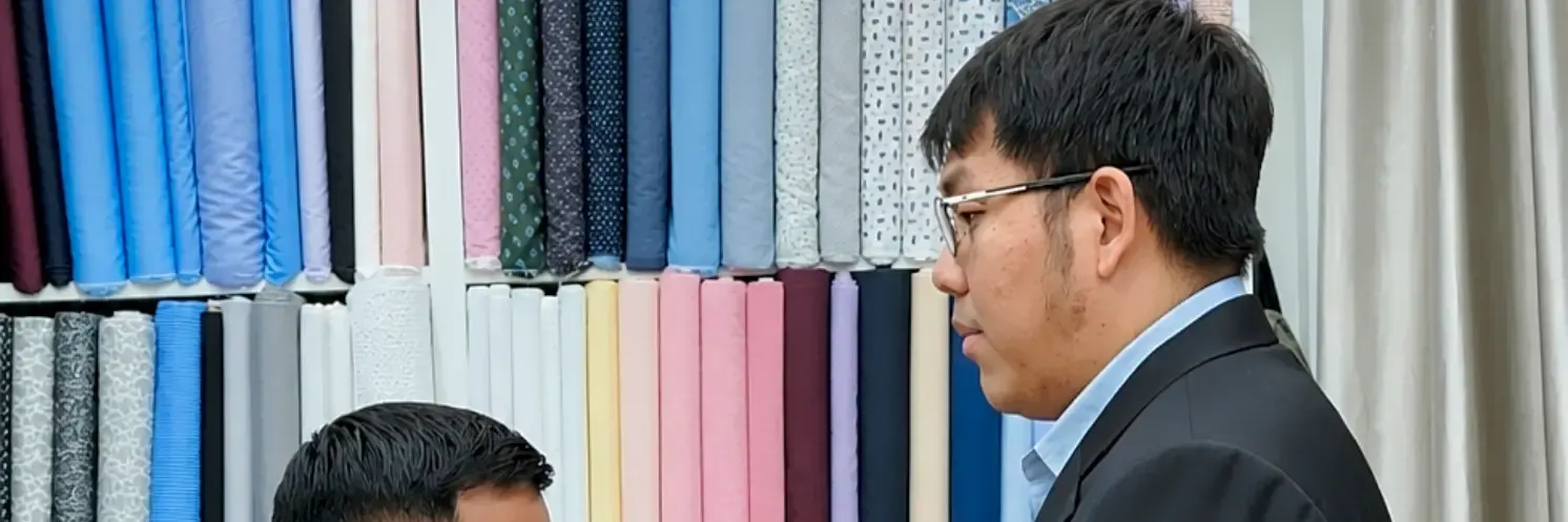 Tailor adjusting a black jacket on a client in front of the fabric wall