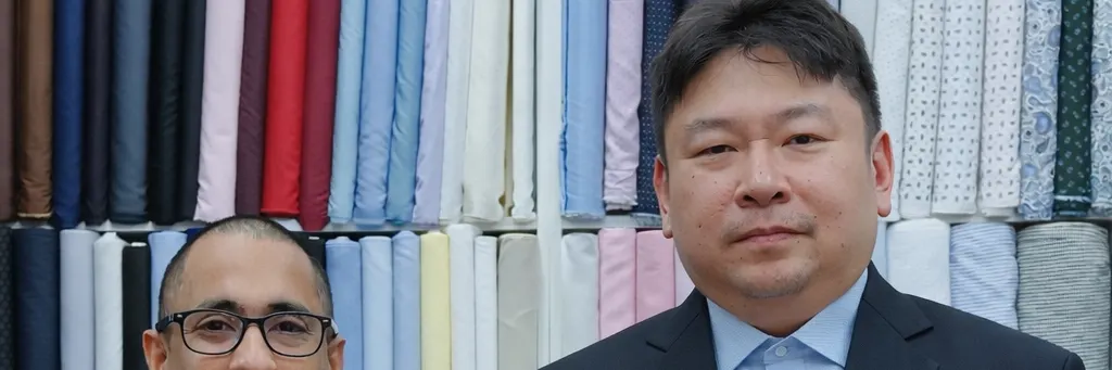 Client in a tailored navy suit standing beside a tailor in front of the fabric wall