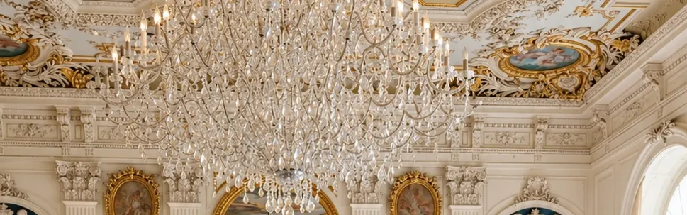 Groom in a white dinner jacket and bride in a lace gown in an ornate hall with a chandelier