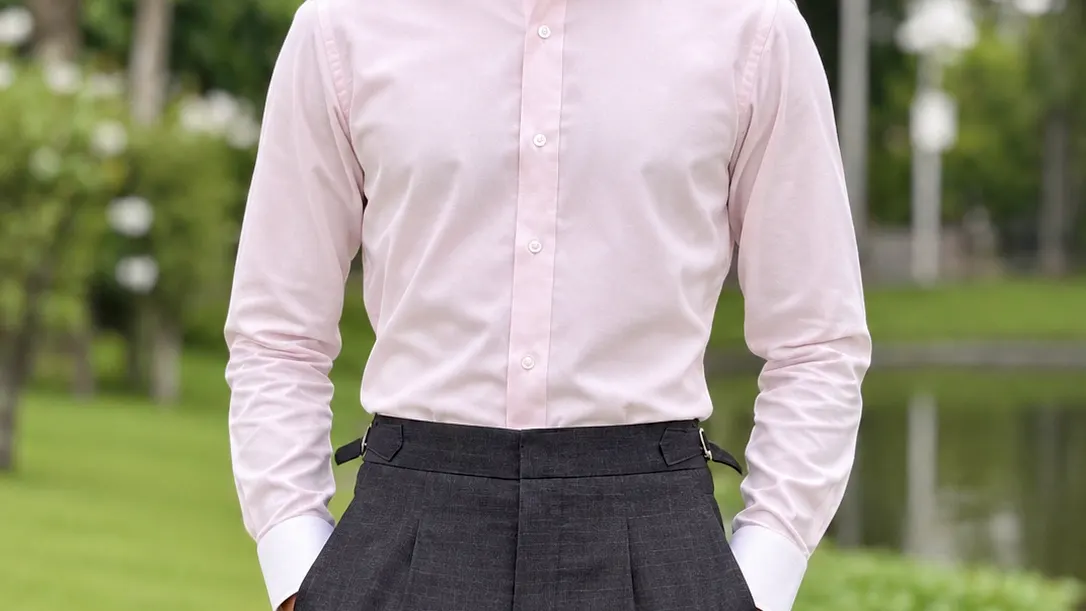Man in a tailored light pink dress shirt and high-waisted grey trousers standing by a pond