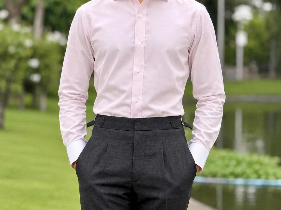 Man in a tailored light pink dress shirt and high-waisted grey trousers standing by a pond