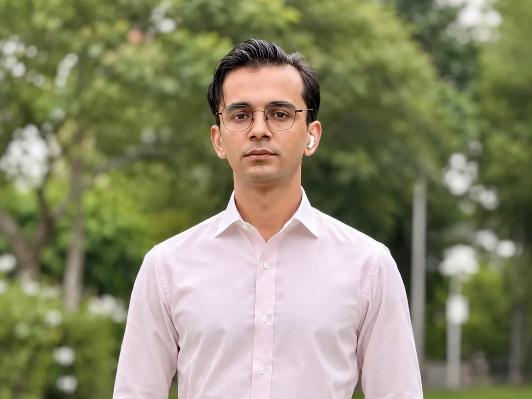Man in a tailored light pink dress shirt and high-waisted grey trousers standing by a pond
