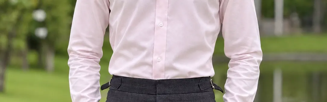 Man in a tailored light pink dress shirt and high-waisted grey trousers standing by a pond