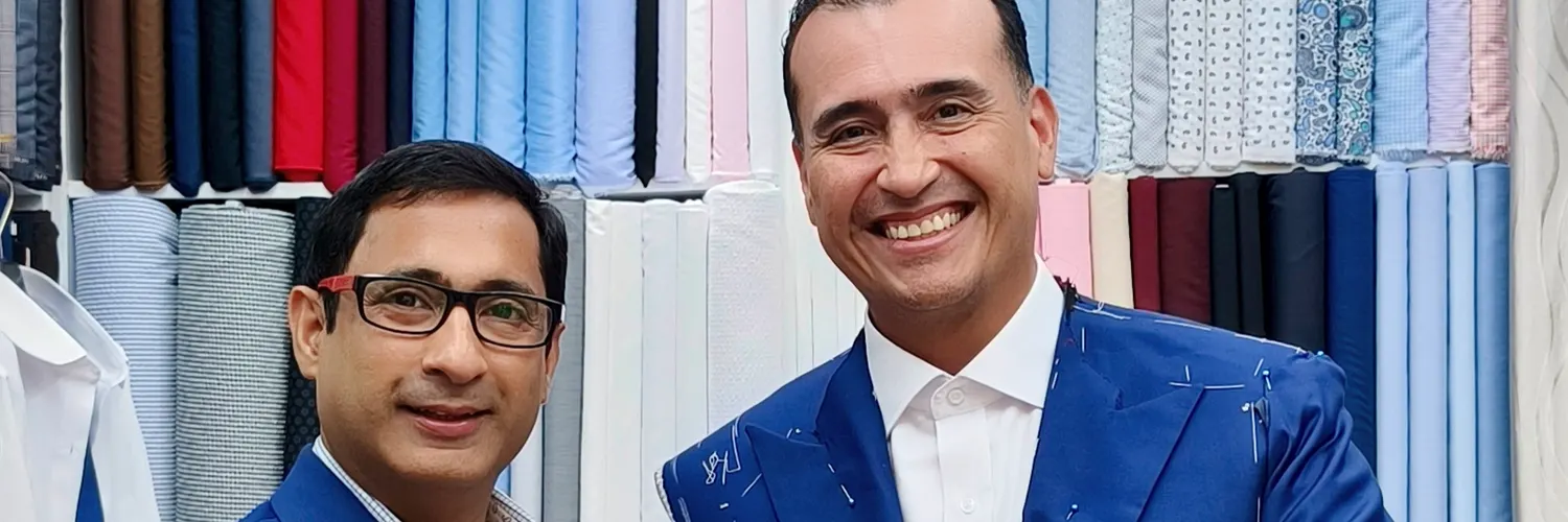Client wearing a blue jacket pinned and chalked for a fitting, next to a tailor in front of the fabric wall