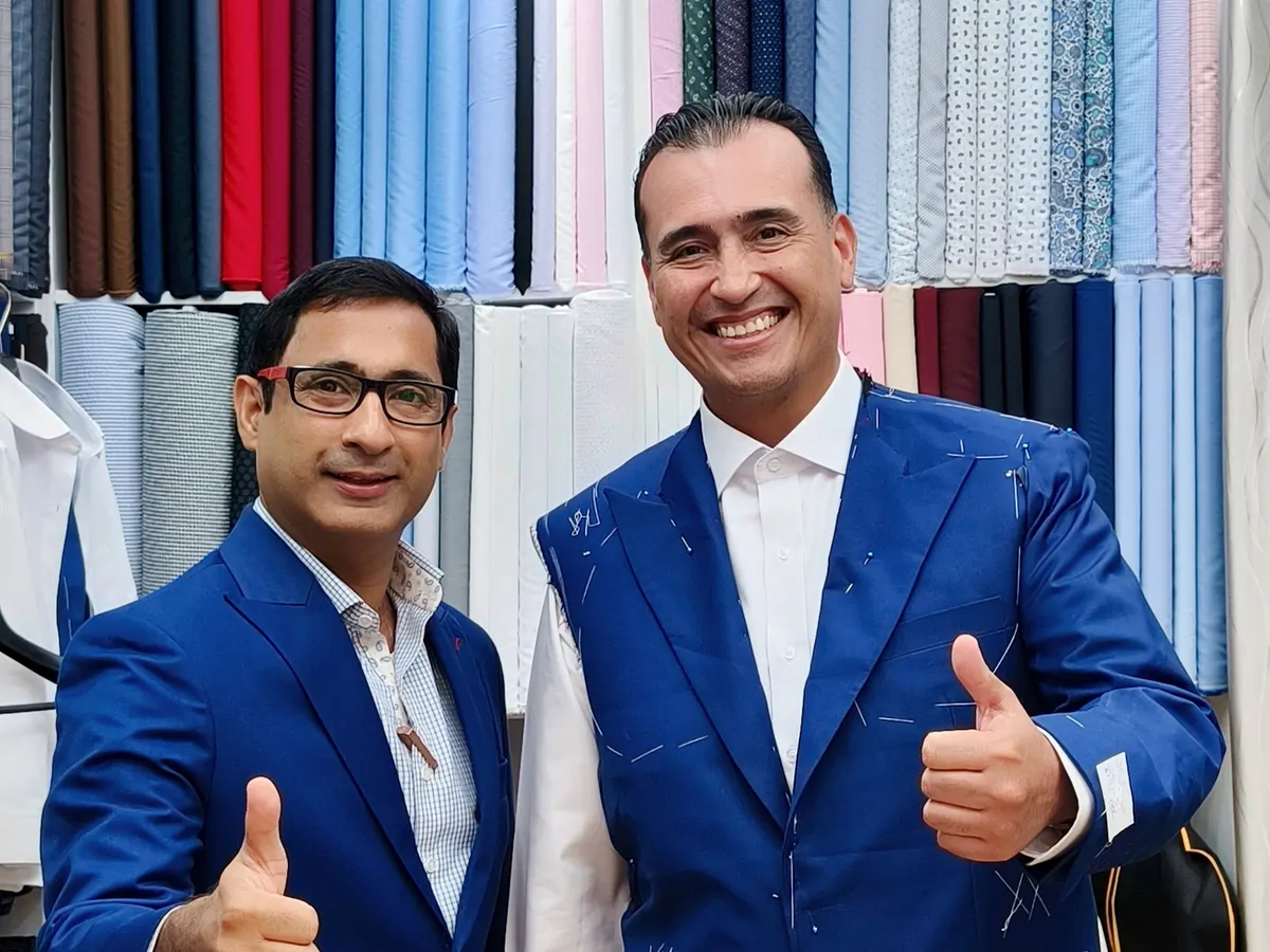 Client wearing a blue jacket pinned and chalked for a fitting, next to a tailor in front of the fabric wall