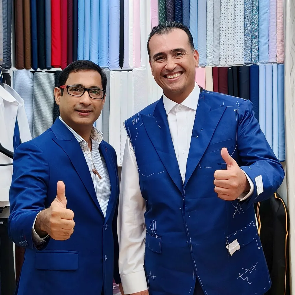 Client wearing a blue jacket pinned and chalked for a fitting, next to a tailor in front of the fabric wall