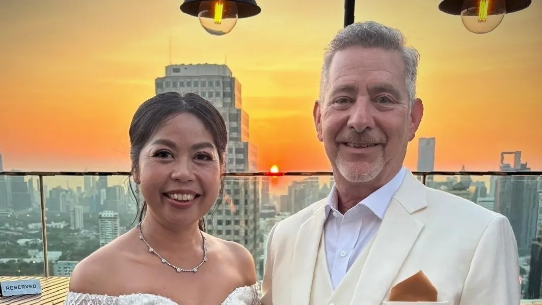 Bride in a feathered white dress and groom in a cream three-piece suit at sunset on a rooftop