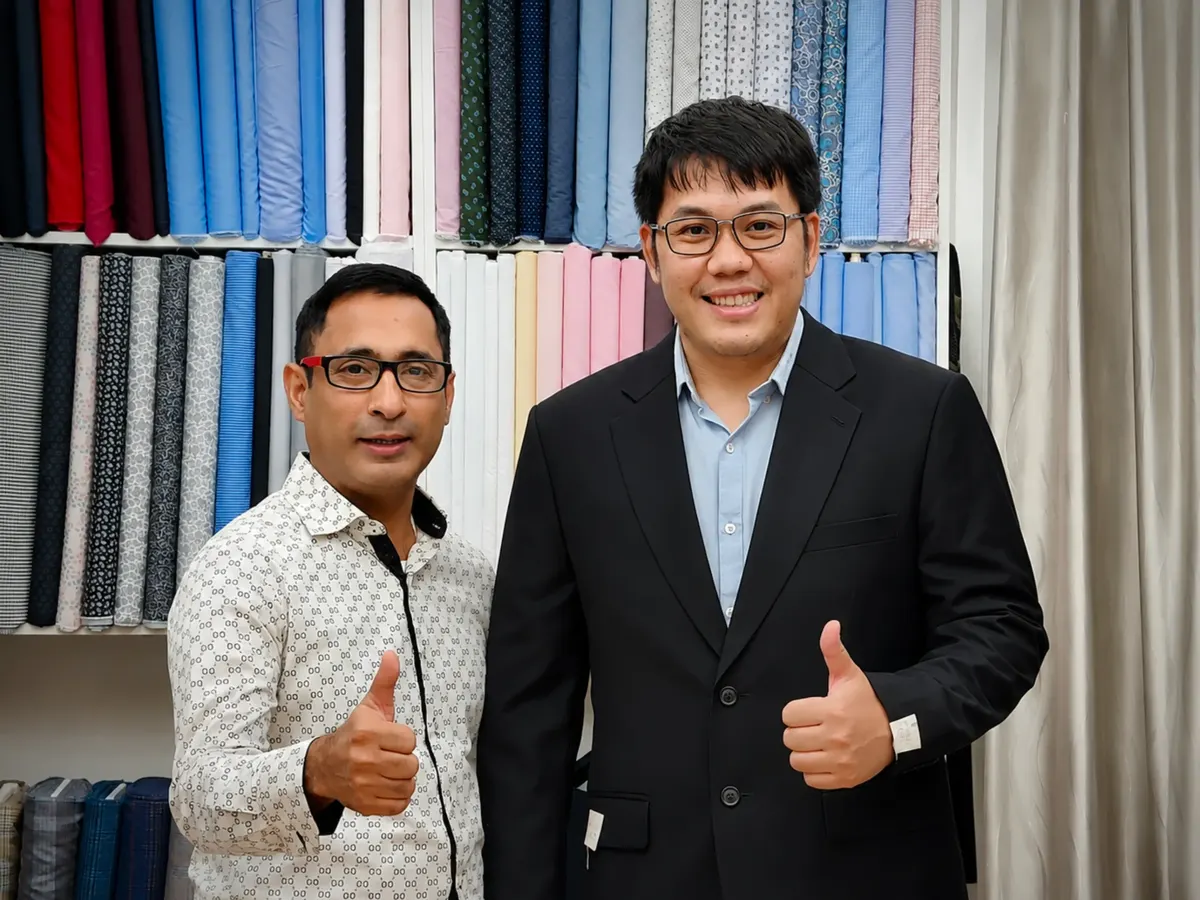 Tailor and a client in a black suit jacket smiling in front of the fabric wall